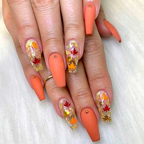 Cute matte orange fall nails combined with clear nail tips which, adorned with fall leaves glitter and gold foil patches.