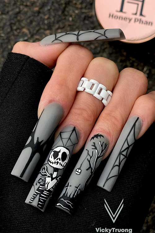 Long matte grey nails with black and white Halloween nail art ideas feature a skeleton, cemetery, cobweb, and bat 