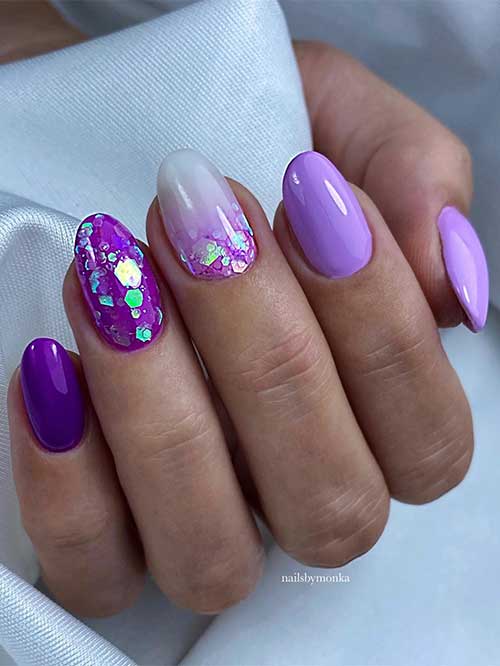 Short different shades of purple nails with glitter and an accent white nail with glitter.