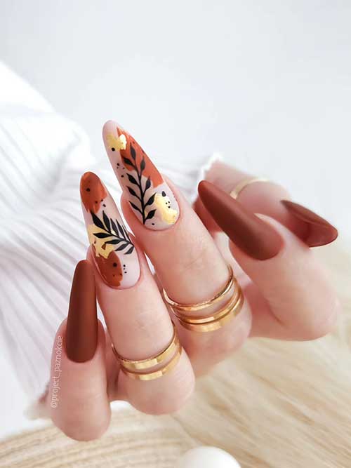 Long almond brown nails with two nude accent nails adorned with brown and gold foil abstract nail art and black leaf nail art