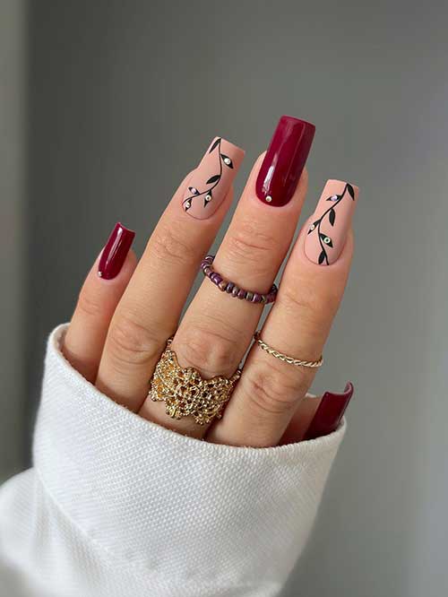 Long square shaped dark red nails with two accent matte nude nails adorned with black leaf nail art and rhinestones