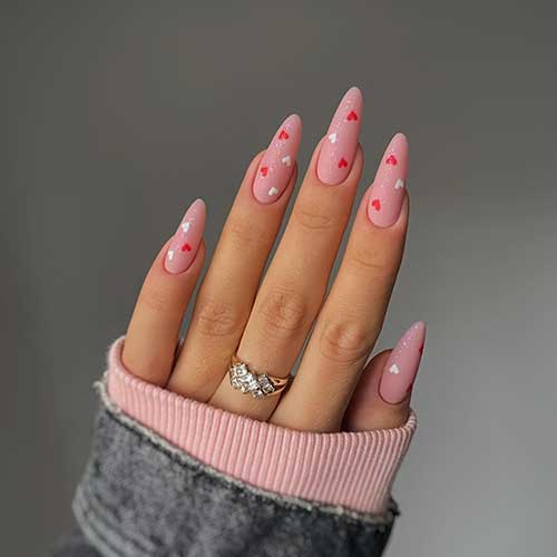 Long almond-shaped matte pink Valentine’s nails with a touch of glitter. And adorned with white and red heart shapes