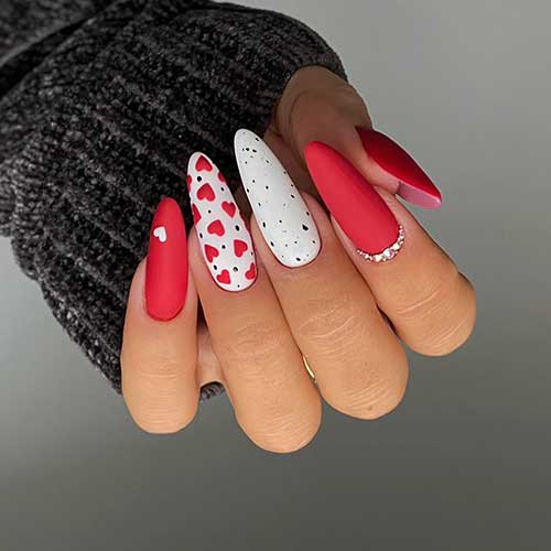 Matte red and white Valentine’s nails are almond-shaped and adorned with black speckles, rhinestones, and red hearts