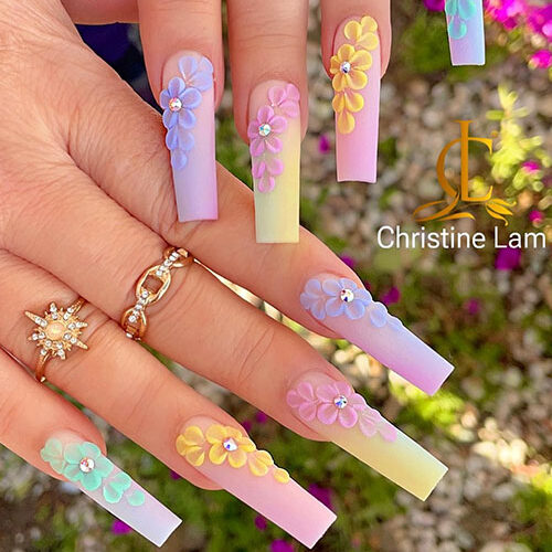 Long matte multicolored nails feature yellow, pink, blue, and mint green colors and are adorned with flowers and rhinestones