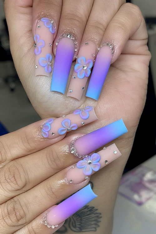Long matte nude purple blue ombre nails with two accent nude nails adorned with prominent purple-blue flowers and crystals
