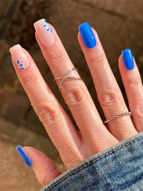 Bold blue nails paired with delicate floral patterns on nude accents create a stylish and sophisticated nail design. Bold blue nails paired with delicate floral patterns on nude accents create a stylish and sophisticated nail design.