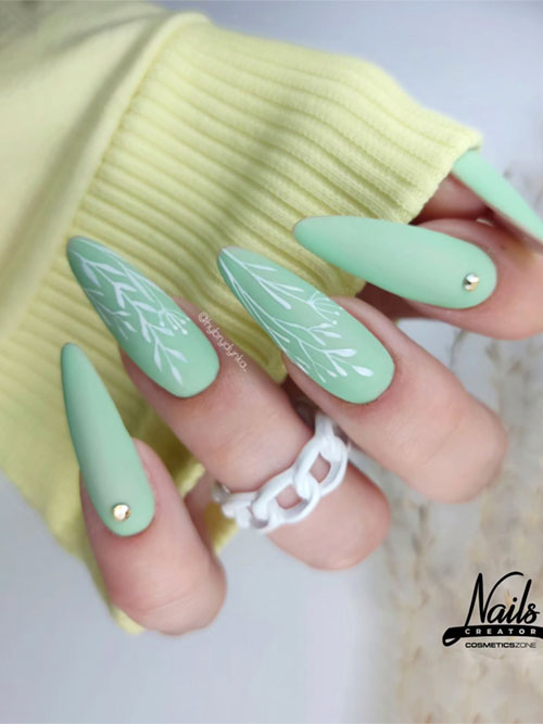 Soft pastel green matte nails with white leaf patterns and gold studs, paired with a white chain-link ring for a fresh spring look. Soft pastel green matte nails with white leaf patterns and gold studs, paired with a white chain-link ring for a fresh spring look.